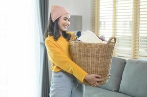 jovem mulher feliz vestindo camisa amarela segurando uma cesta cheia de roupas em casa, conceito de lavanderia foto