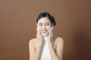 uma jovem com rosto bonito sorrindo, lavando o rosto sobre fundo marrom, conceito de cuidados com a pele de beleza foto