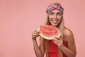 retrato de mulher loira feliz com cabelo comprido, segurando o pedaço de melancia, olhando de lado e sorrindo amplamente, posando sobre fundo rosa foto