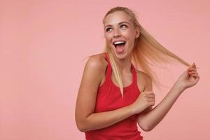 jovem loira feliz de pé sobre fundo rosa com a boca aberta, olhando para cima com um sorriso largo e puxando o cabelo, conceito de emoções positivas foto