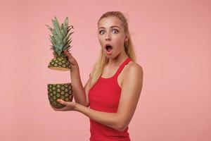 retrato de jovem atraente espantada com duas partes de abacaxi, olhando para a câmera com cara de surpresa, de pé sobre fundo rosa foto