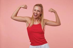 jovem bonita alegre com penteado casual, mostrando as mãos esportivas, sorrindo amplamente, mostrando a língua e piscando para a câmera, isolada sobre fundo rosa foto