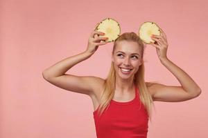 alegre mulher jovem e bonita com maquiagem natural e roupas casuais, olhando de lado com um sorriso largo, levantando as mãos com pedaços de abacaxi na cabeça foto