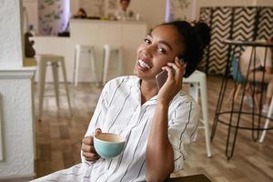 mulher de pele escura atraente feliz com penteado de coque falando no telefone com um amigo enquanto bebe café no café, olhando para a câmera alegremente foto