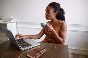 foto interna de linda mulher encaracolada de pele escura posando sobre o espaço de trabalho com cara séria, bebendo café enquanto digita notas em seu laptop