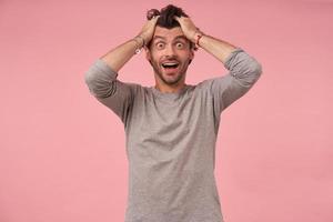 foto de estúdio de belo homem barbudo espantado com corte de cabelo na moda vestindo roupas casuais, posando sobre fundo rosa com as mãos na cabeça, olhando para a câmera com rosto atordoado e boca aberta