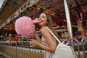 feliz jovem morena atraente com cabelos compridos andando pelo parque de diversões, usando vestido leve de verão e mochila branca, puxando um pedaço de algodão doce com os dentes foto