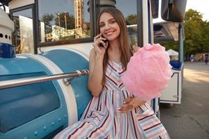 jovem adorável mulher de cabelos compridos em vestido romântico sentado no parque de diversões e apoiando-se no trem a vapor falso, tendo uma conversa agradável no telefone e sorrindo alegremente para a câmera com algodão doce na mão foto