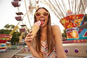 jovem mulher bonita positiva com longos cabelos castanhos posando sobre parque de diversões em dia quente de verão, lambendo sorvete com sorriso largo, usando óculos escuros e vestido romântico leve foto
