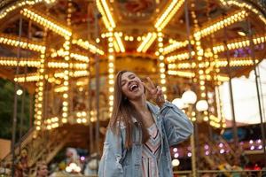 alegre jovem atraente com cabelo comprido castanho posando sobre carrossel com casaco jeans, levantando a mão com gesto de vitória, sorrindo com a boca aberta e fechando os olhos foto