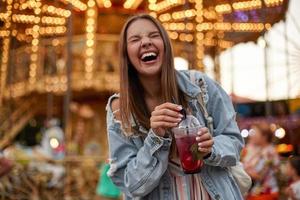 alegre mulher jovem e bonita com cabelo castanho em roupas casuais, bebendo limonada enquanto caminhava no parque de diversões, rindo alto com os olhos fechados e franzindo foto