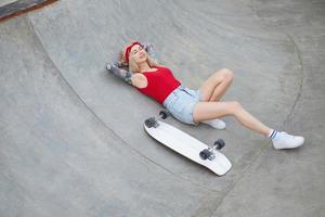 jovem tatuada linda mulher de cabelos compridos na faixa de cabeça posando sobre skate park, deitada no chão de concreto com os olhos fechados e mantendo as mãos debaixo da cabeça foto