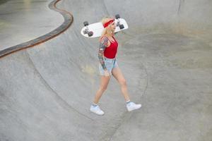 foto ao ar livre da jovem loira tatuada de cabelos compridos linda mulher andando sobre o parque de skate em roupas casuais, mantendo a prancha de skate nos ombros