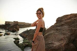 jovem mulher bonita em roupas romantice e óculos de sol vintage posando sobre a vista à beira-mar, mantendo a mão no pescoço e sorrindo sinceramente para a câmera foto