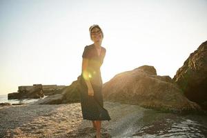 fêmea jovem positiva com penteado casual em pé sobre a baía do mar em dia ensolarado e quente, usando vestido de verão romântico e sorrindo sinceramente para a câmera foto