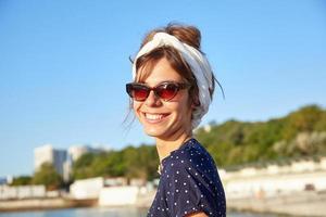 close-up de feliz linda mulher de cabelos escuros na faixa de cabeça e óculos de sol olhando para a câmera alegremente, posando ao ar livre em dia quente e ensolarado foto