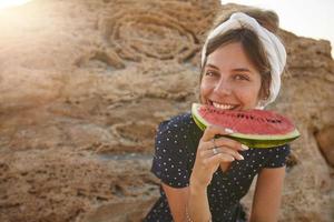 foto ensolarada ao ar livre da bela jovem morena sentada sobre uma grande pedra amarela e comendo melancia com a mão, estando de bom humor e sorrindo sinceramente