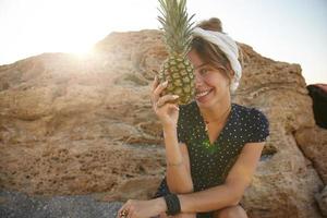 foto ensolarada ao ar livre de encantadora jovem morena com vestido de verão e bandana sentado sobre pedra amarela, segurando abacaxi fresco na mão e sorrindo para a câmera alegremente