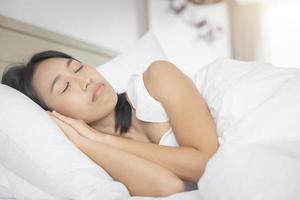 jovem mulher asiática dormindo na cama no quarto. ela está deitada de lado e relaxa com os olhos fechados foto