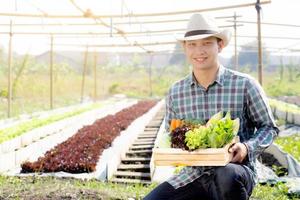 retrato jovem asiático sorrindo colheita e pegando horta orgânica fresca na cesta na fazenda hidropônica, agricultura e cultivo para alimentação saudável e conceito de negócios. foto