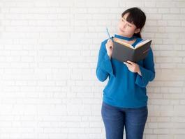 linda mulher asiática sorrindo em pé pensando e escrevendo caderno sobre fundo branco de cimento concreto em casa, lição de casa de menina no conceito de livro, educação e estilo de vida. foto