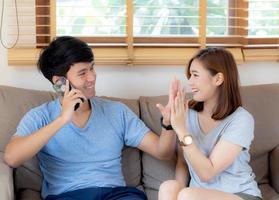 belo jovem casal asiático, homem e mulher, gesticulam cinco juntos enquanto homem falando telefone móvel inteligente no sofá, festa de família, aproveite e animado com feliz no sofá, conceito de estilo de vida. foto