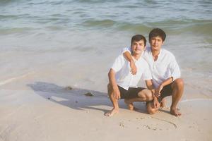 jovem casal asiático gay sorrindo romântico desenho coração forma juntos na areia em férias, homossexual feliz e divertido abraço com amor sentado na areia na praia em viagens de verão, conceito legal lgbt. foto