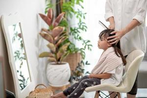 mãe asiática cortando cabelo para a filha na sala de estar em casa enquanto fica em casa segura do coronavírus covid-19 durante o bloqueio. conceito de auto-quarentena e distanciamento social. foto