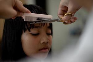 mãe asiática cortando cabelo para a filha na sala de estar em casa enquanto fica em casa segura do coronavírus covid-19 durante o bloqueio. conceito de auto-quarentena e distanciamento social. foto