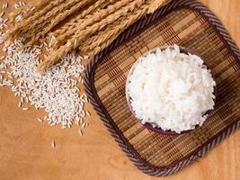 arroz cozido em tigela com grão de arroz cru e planta de arroz seco no fundo da mesa de madeira. foto