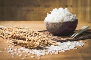 arroz cozido em tigela com grão de arroz cru e planta de arroz seco no fundo da mesa de madeira. foto