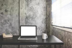 laptop com tela branca em branco, xícara de café branca e telefone na mesa foto