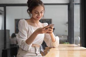 jovem asiática usando telefone inteligente no café. mulher sorridente usando smartphone em casa, mensagens ou navegando nas redes sociais enquanto relaxa. foto