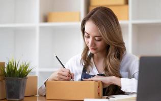 jovem linda e feliz empresária asiática dona de sme on-line usando laptop receber pedido do cliente com embalagem de caixa de encomendas em seu escritório em casa de inicialização, vendedor de negócios on-line e entrega foto