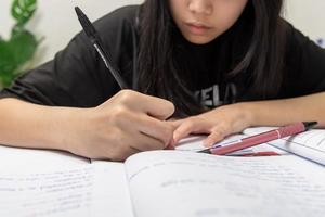 aluna asiática está escrevendo lição de casa e lendo livro na mesa foto
