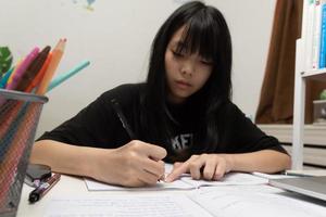 aluna asiática está escrevendo lição de casa e lendo livro na mesa foto