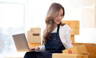 retrato, empresário, jovem mulher asiática, negócios freelance, negócios sme, compras on-line, trabalhando no computador portátil com caixa de encomendas na mesa do escritório em casa, negócios on-line e conceito de entrega. foto