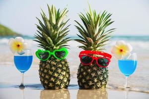 abacaxis de casal fresco com óculos de sol e copos de coquetel na praia de areia limpa com fundo de ondas do mar - frutas frescas e bebida com conceito de fundo de férias de sol de areia do mar foto