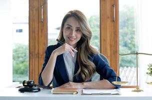 retrato, atraente e encantadora jovem empresária asiática ou gerente executiva feminina senta-se em sua mesa de escritório na moderna sala de escritório brilhante. foto