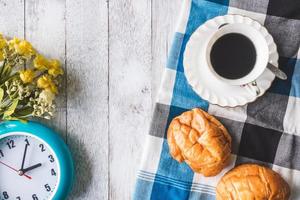 vista superior da xícara de café com pão, toalha de mesa, flores e relógio no fundo da mesa de madeira foto