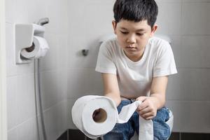 menino asiático sentado no vaso sanitário segurando papel de seda - conceito de problema de saúde foto