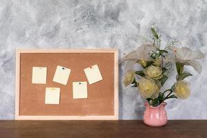 papéis em branco pin up na placa de cortiça e vaso de flores sobre a mesa de madeira com plano de fundo texturizado. foto