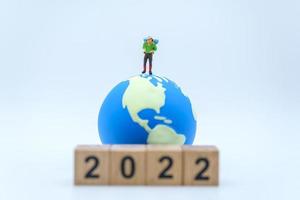 2022 ano novo e conceito de viagens. closeup de figuras em miniatura de viajante masculino com pé de mochila com pilha de bloco de número de madeira e mini bola mundial em fundo branco.