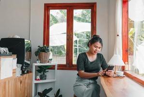 mulher asiática com um lindo sorriso assistindo no celular durante o descanso em uma cafeteria, feliz mulher tailandesa sentada no balcão de bar de madeira tomando café relaxando no café durante o tempo livre foto