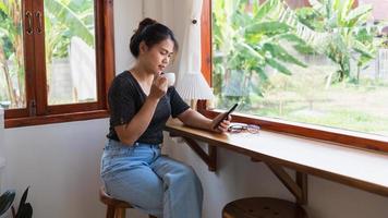 mulher asiática com um lindo sorriso assistindo no celular durante o descanso em uma cafeteria, feliz mulher tailandesa sentada no balcão de bar de madeira tomando café relaxando no café durante o tempo livre