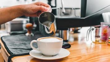 o barista profissional despeja o leite na caneca de aço inoxidável na xícara de café quente para fazer um cappuccino ou café com leite na cafeteria. como fazer café capuccino foto
