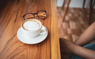 xícara de café cappuccino com café com leite e copos de uma mulher em um balcão de bar de madeira em um café ensolarado pela manhã. foto