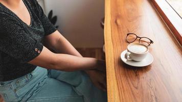 xícara de café cappuccino com café com leite e copos de uma mulher em um balcão de bar de madeira em um café ensolarado pela manhã. foto