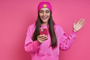 mulher jovem e bonita na camisa com capuz e chapéu funky segurando telefone inteligente contra fundo rosa foto