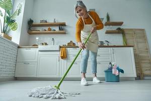 comprimento total da mulher sênior, limpando o chão com esfregão em pé na cozinha doméstica foto